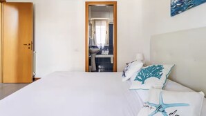 A bedroom with a bed, a wooden door, a bathroom visible through an open door, and a painting on the wall.