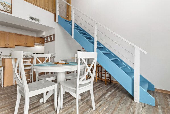 A modern kitchen with white cabinetry, a blue staircase, and a dining area with white chairs and a round table. - The Sunset is Calling at Shells Sunset Cove (Avon)