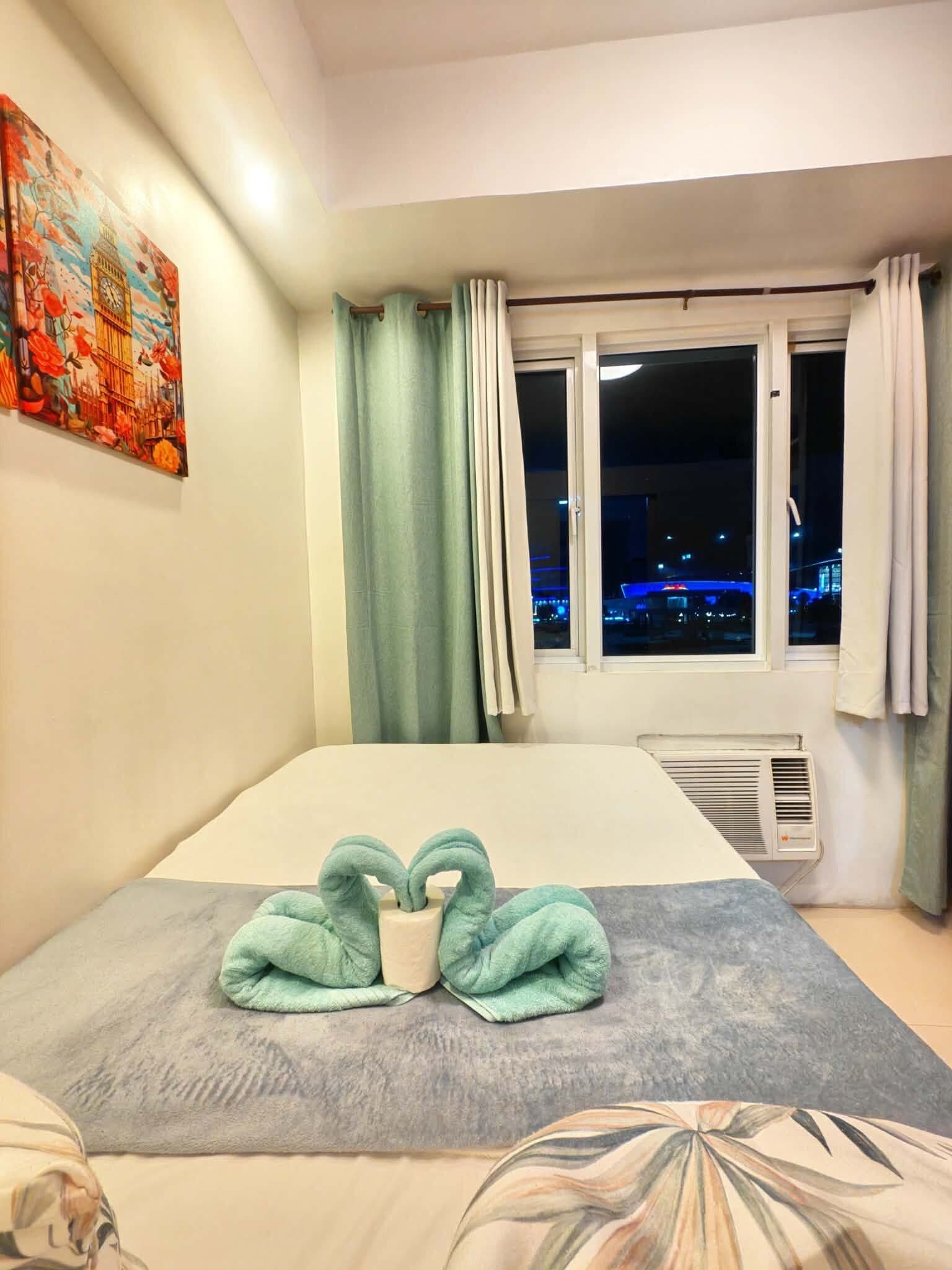 A hotel room with a bed, a window with curtains, a wall-mounted air conditioner, and a painting.