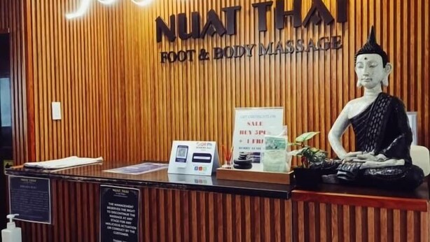 Un comptoir d’accueil avec un panneau en bois en arrière-plan, une statue de Bouddha et une enseigne indiquant « NUAT THAI FOOT & BODY MASSAGE ».