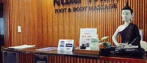 Un comptoir d’accueil avec un panneau en bois en arrière-plan, une statue de Bouddha et une enseigne indiquant « NUAT THAI FOOT & BODY MASSAGE ».