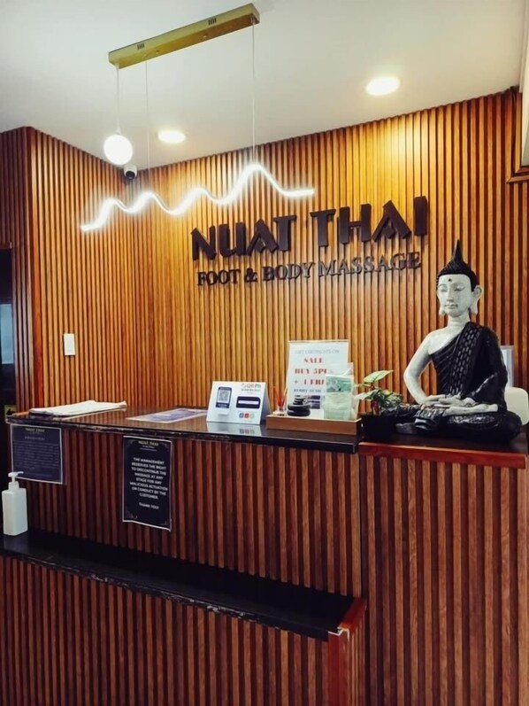 En receptionsdisk med en bakgrund av träpaneler, en Buddha-staty och en skylt med texten ”NUAT THAI FOOT & BODY MASSAGE”.
