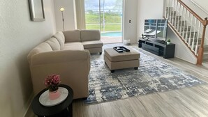 Ein modernes Wohnzimmer mit einem Ecksofa, einem kleinen runden Tisch mit einer rosa Vase, einem schwarzen Hocker, einem grauen und weißen Teppich mit Muster, einem Deckenventilator, einem Fernseher auf einem schwarzen Standfuß und einer Treppe mit weißen Geländern.