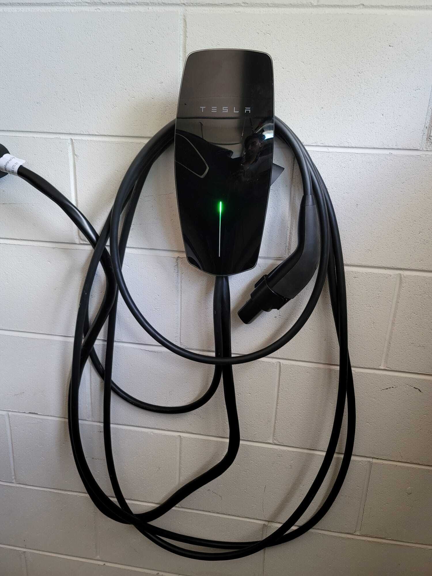 Une borne de recharge Tesla dotée d’un appareil de recharge noir et d’un indicateur lumineux vert, le tout relié par plusieurs câbles noirs.