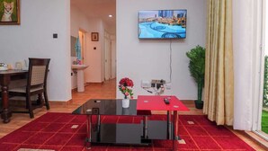 Un salon moderne avec une table basse en verre, une petite table d’appoint rouge, un téléviseur fixé au mur et une vue sur le paysage urbain.