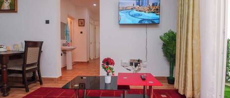 A modern living room with a glass coffee table, a red accent table, a TV mounted on the wall, and a view of a cityscape.