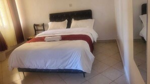 Un lit bien fait, avec du linge de lit blanc rehaussé d’une touche de rouge, une petite table de chevet et une chaise.