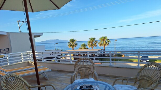 Una terraza con sillas de mimbre y una mesa de vidrio, con vistas a una playa con palmeras y un coche estacionado cerca.