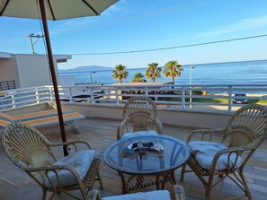 Une terrasse dotée de chaises en osier et d’une table en verre, offrant une vue sur une plage bordée de palmiers et sur une voiture garée à proximité.