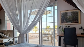 Un dormitorio con cama, cortinas blancas, escritorio, silla y un cuadro en la pared.