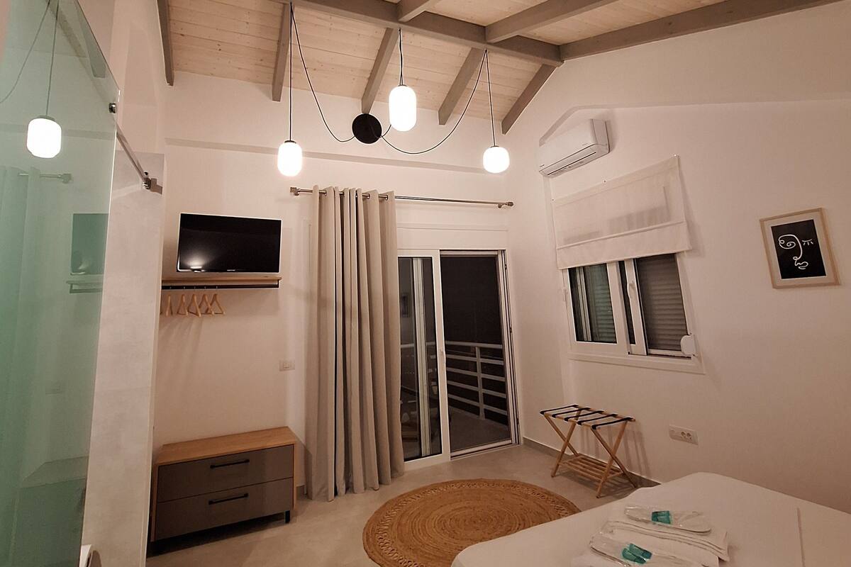 A bedroom with a wooden ceiling, a flat-screen TV, a sliding glass door, a bed with neatly folded towels, and a small wooden bench.