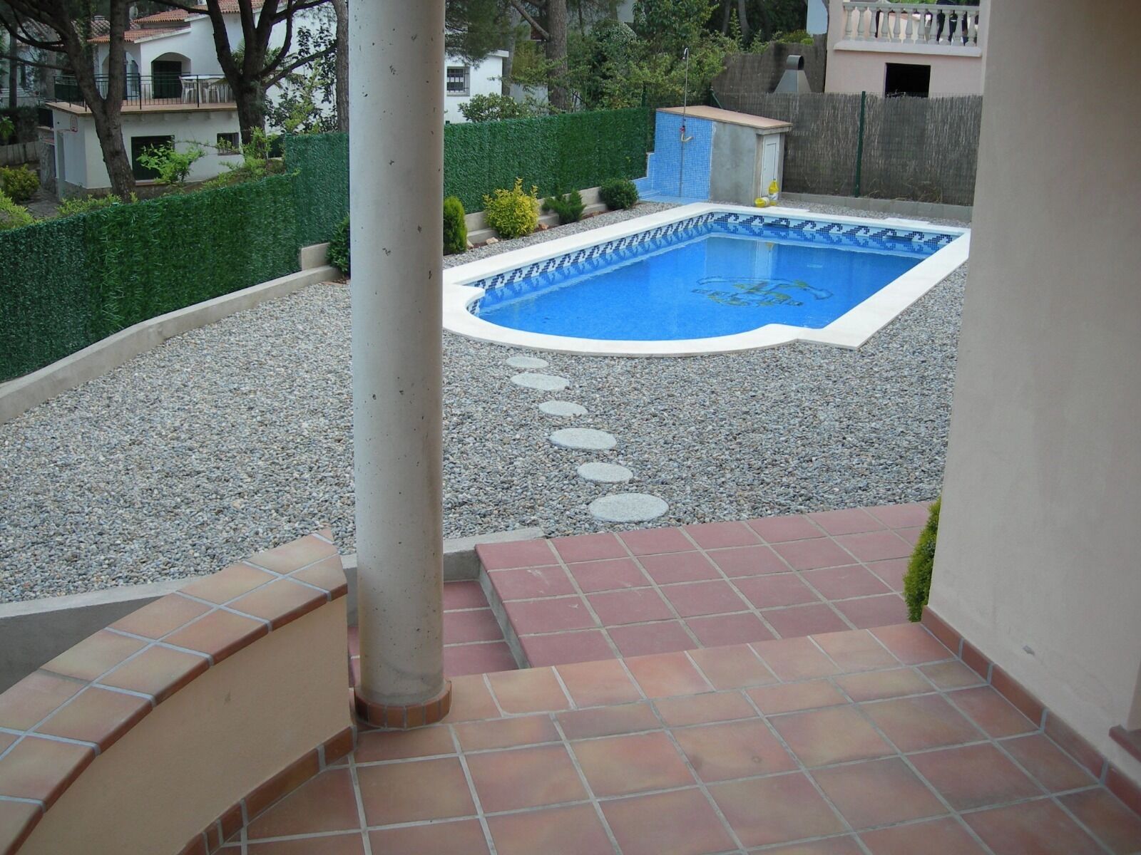 En swimmingpool med en kant av kakel, omgiven av ett område med grus och stenplattor som man kan gå på, samt en uteplats med kakel.