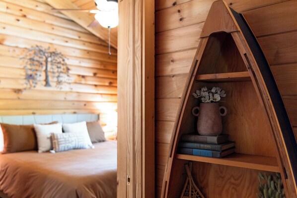 Un interior de madera con una cama, un estante con libros y un florero, y una pared con un diseño de árbol.