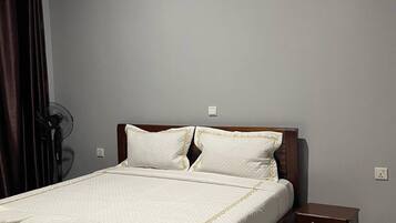 A neatly made bed with a wooden frame, a bedside table, and a wall-mounted fan.