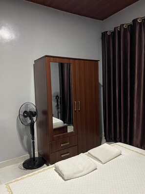Ein Schlafzimmer mit einem Holzschrank, einem Standventilator, einem Bett mit weißer Bettwäsche und dunklen Vorhängen.