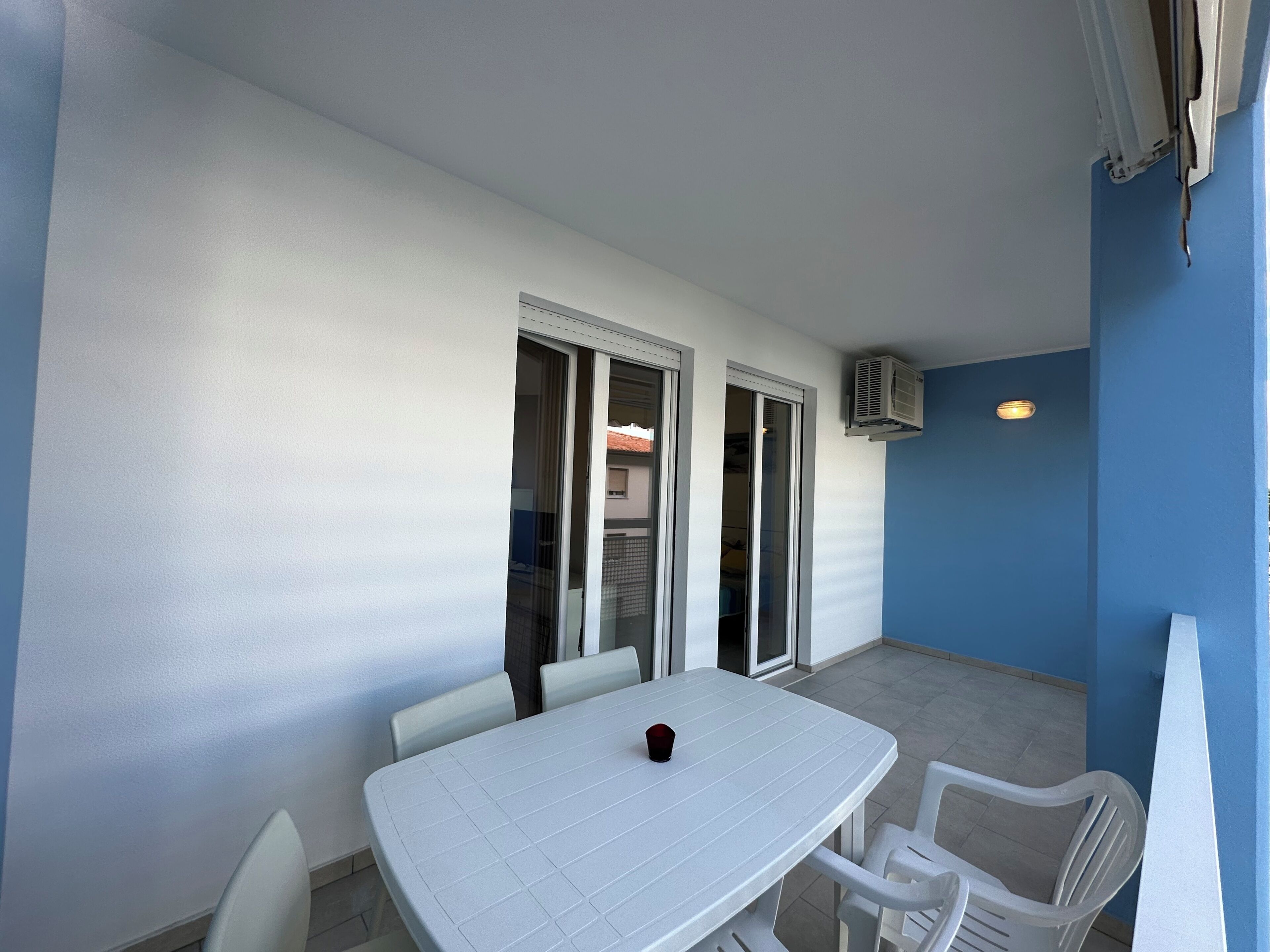 Un balcon avec une table et des chaises blanches, une porte-fenêtre coulissante et un appareil de climatisation.