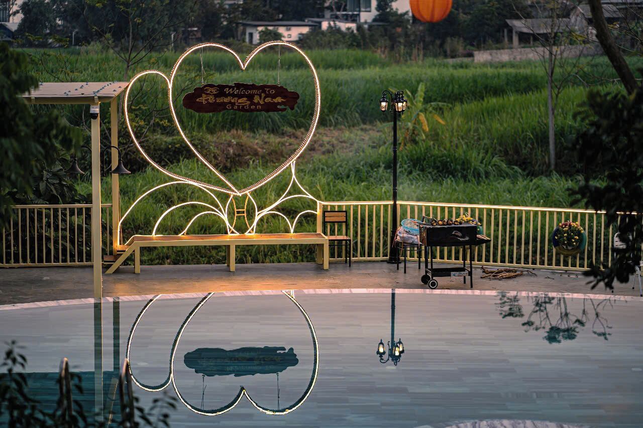 A heart-shaped sign with a bench and a table with flowers, surrounded by a fence and greenery.