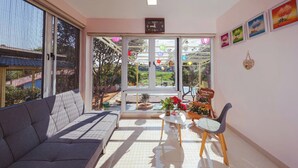 Ein modernes Wohnzimmer mit einem grauen Sofa, einem kleinen Tisch mit Topfpflanzen und einem Sessel. Große Fenster bieten Blick auf einen Garten und ein Gebäude.