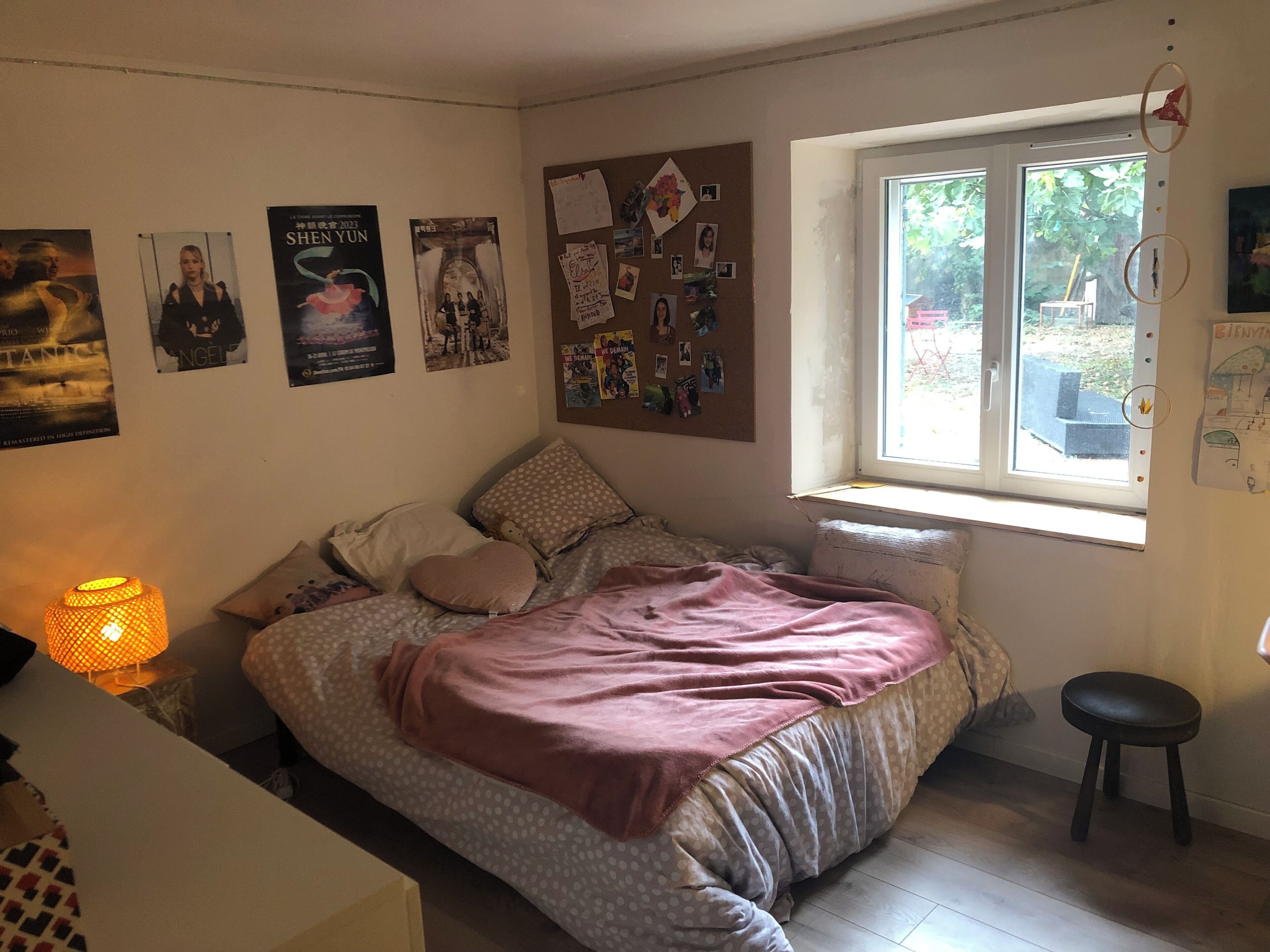 Een slaapkamer met een bed, een raam en wanddecoraties, waaronder posters en een prikbord.