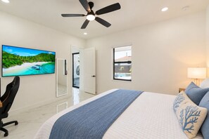 Ein Schlafzimmer mit Bett, Deckenventilator, Fernseher, Schreibtisch und Fenster mit Blick auf einen Pool und ein Gebäude.