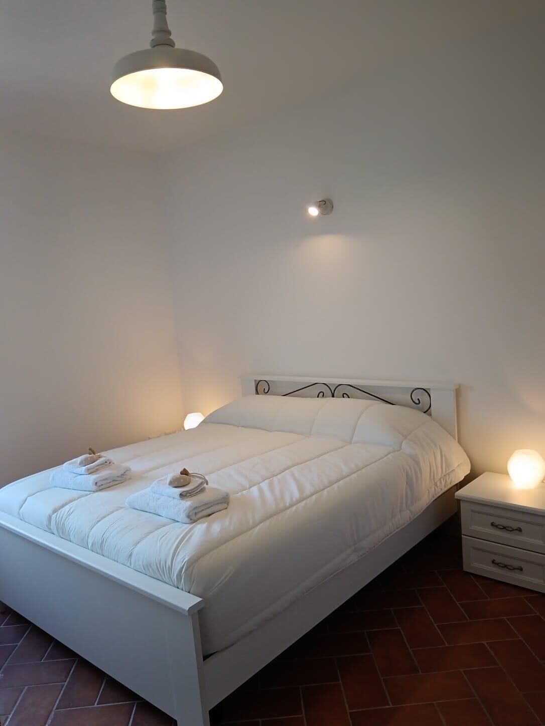 A neatly made bed with white bedding and two towels, a bedside table with a lamp, and a ceiling light fixture.