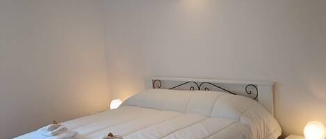 A neatly made bed with white bedding and two towels, a bedside table with a lamp, and a ceiling light fixture.