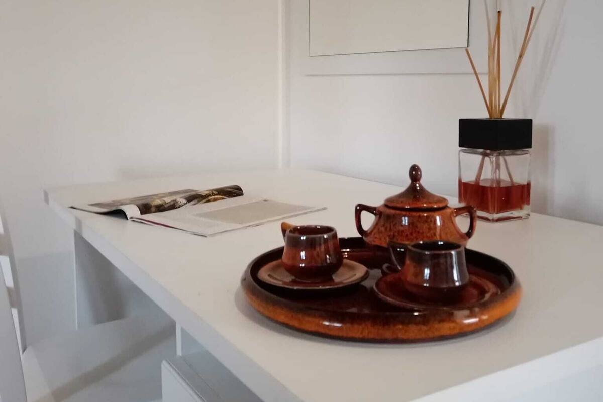 A white desk with a tray holding a teapot and cups, a magazine, and a small vase with reed sticks.
