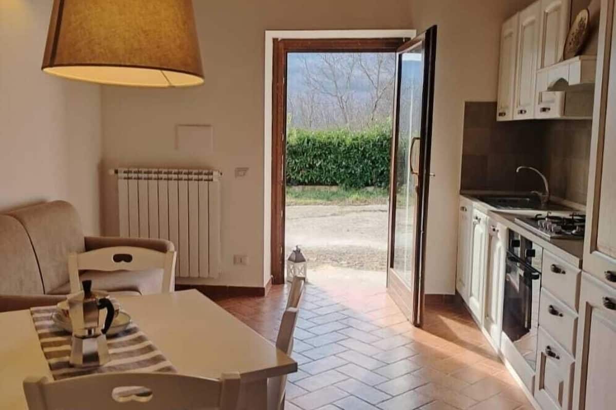 A cozy kitchen with a dining area, wooden ceiling beams, and a view through an open door to the outside.