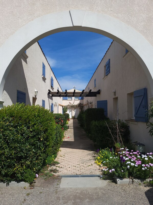 A narrow alleyway with a white archway, leading to a building with blue shutters and a tiled roof.