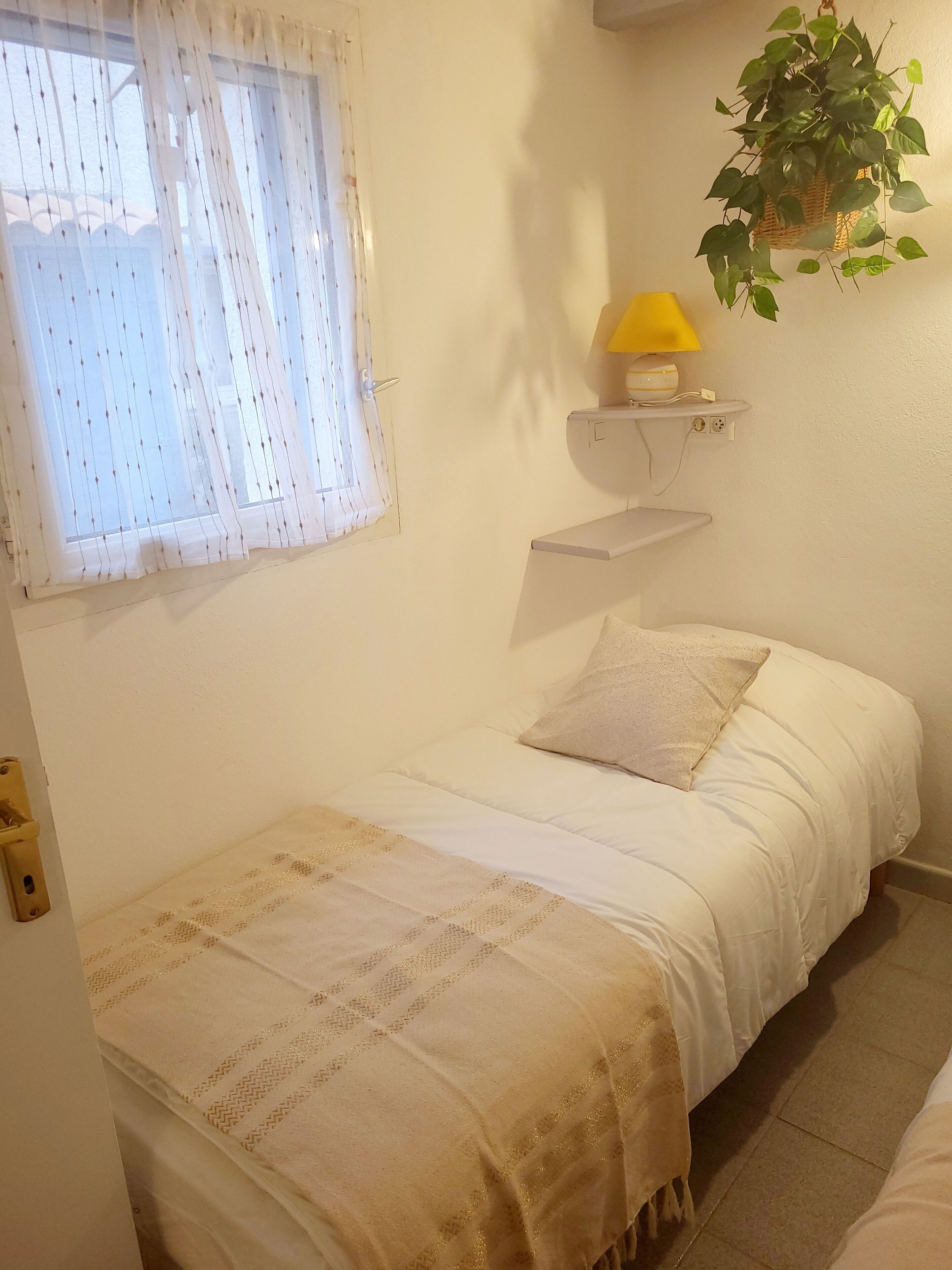 Un dormitori amb un llit, una finestra amb cortines lleugeres, una làmpada de nit i una planta penjant.