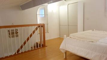 Quarto com piso de madeira, cama branca, corrimão branco e guarda-roupa com portas deslizantes.