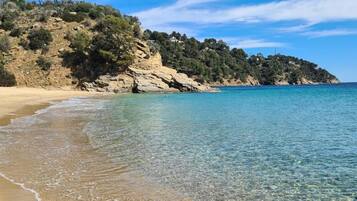 Uma praia com águas cristalinas, areal arenoso e uma colina rochosa coberta de vegetação.