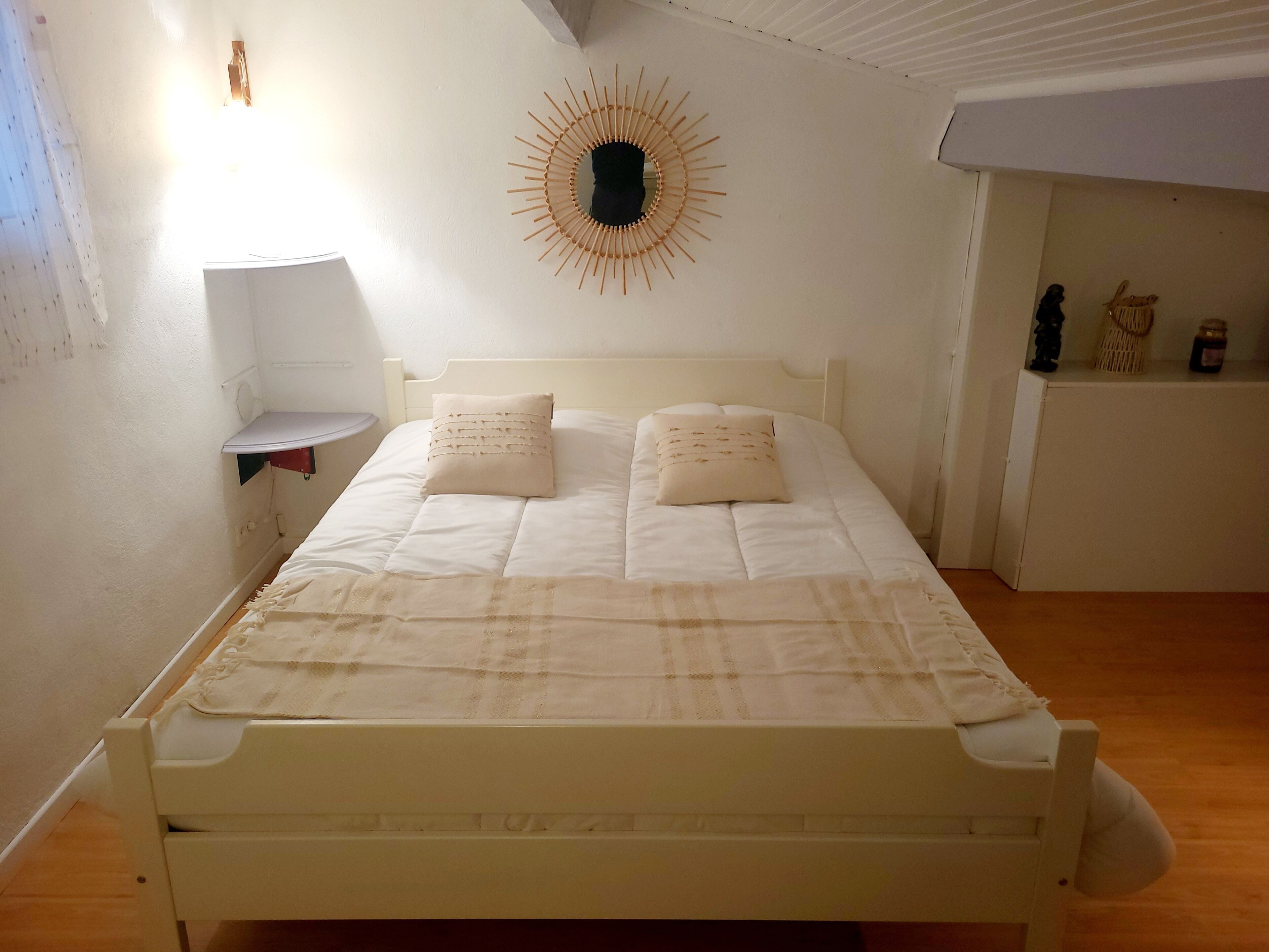 A bedroom with a bed, two pillows, a wall-mounted sun decoration, a small bedside table, and a shelf with decorative items.