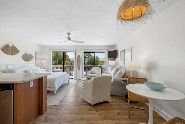 A modern hotel room with a kitchenette, a bed, a seating area with a sofa and chairs, a dining table with a bowl, and a ceiling fan. - The Mini Pearl 30A beach studio - Dog Friendly! (Santa Rosa Beach)