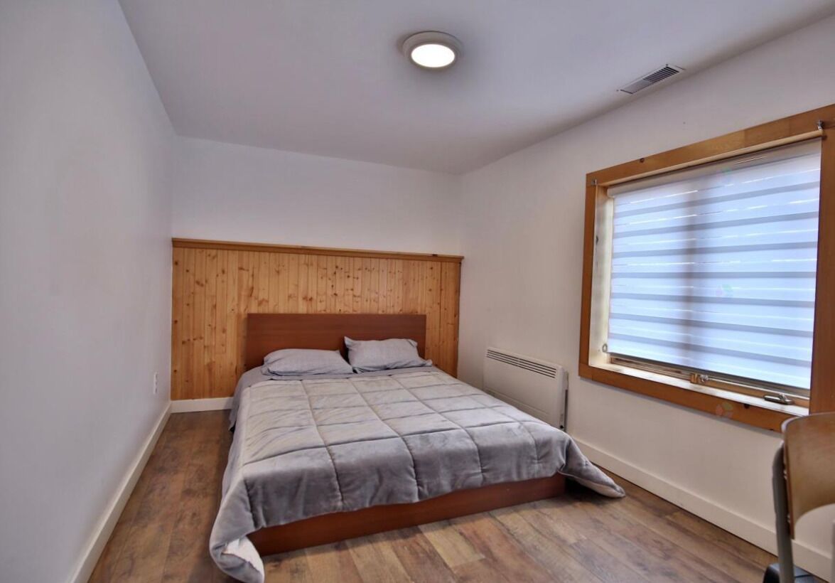 Un dormitorio con cabecera de madera, cama con ropa de cama gris, ventana con persianas y radiador debajo de la ventana.