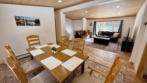 Ein Essbereich mit Holztisch und Stühlen, einer kleinen Pflanze auf dem Tisch und Blick auf ein Wohnzimmer mit Sofa und Fernseher.