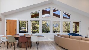 A modern interior with a dining area, a sofa, and large windows offering a view of trees and mountains. - 3 King Suites Near Alyeska | Sauna & Mountain Views (Girdwood)