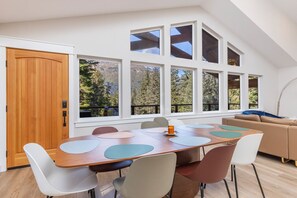 A modern dining area with a wooden table, white and brown chairs, and large windows offering a view of trees and mountains. - 3 King Suites Near Alyeska | Sauna & Mountain Views (Girdwood)