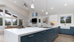 A modern kitchen with a large island, dark blue cabinets, and white countertops. There are multiple windows providing natural light and views of the outdoors. - 3 King Suites Near Alyeska | Sauna & Mountain Views (Girdwood)