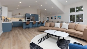 A modern kitchen with blue cabinets, a central island with bar stools, a dining area with a sofa and coffee table, and a large window with a view of trees. - 3 King Suites Near Alyeska | Sauna & Mountain Views (Girdwood)
