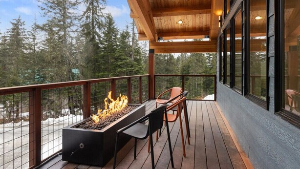 A wooden deck with a fire pit, two chairs, and a view of a forest. - 3 King Suites Near Alyeska | Sauna & Mountain Views (Girdwood)