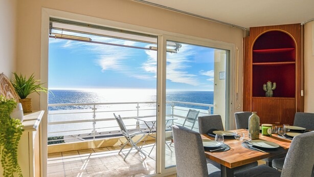 A balcony with a view of the sea, a dining table set with plates and glasses, and a cactus in a wooden display.