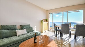 A modern living room with a green sofa, a wooden coffee table with a bottle and glasses, and a dining area with chairs and a table.