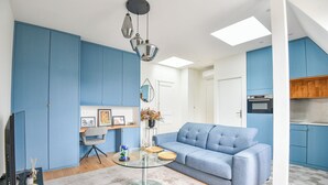A modern living room with a blue sofa, a glass coffee table, and a kitchen area with blue cabinets. - Bail mobilité - Appart climatisé - Arc de Triomphe (Paris)