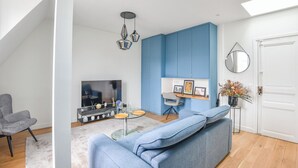 A modern living room with a blue accent wall, a grey sofa, a glass coffee table, and a TV. - Bail mobilité - Appart climatisé - Arc de Triomphe (Paris)