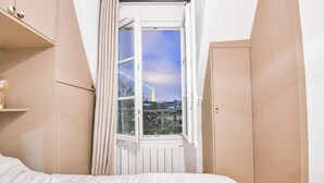 A bedroom with a bed, a window with curtains, a radiator, and a view of a cityscape including a tower. - Bail mobilité - Appart climatisé - Arc de Triomphe (Paris)