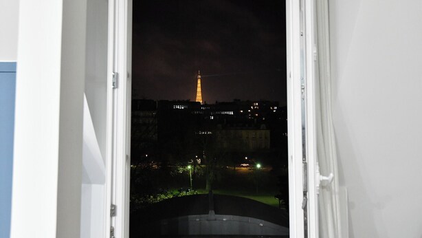 A view through an open door of a building with a nighttime cityscape, including a lit tower resembling the Eiffel Tower. - Bail mobilité - Appart climatisé - Arc de Triomphe (Paris)