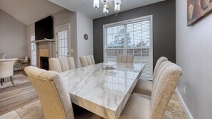 A dining room with a marble table and beige upholstered chairs, a fireplace, and a large window with blinds.