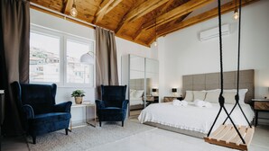 Ein modernes Schlafzimmer mit einem großen Bett, zwei blauen Sesseln, einer Hängeschaukel und Blick auf Gebäude durch ein Fenster.