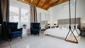 A modern bedroom with a large bed, two blue armchairs, a hanging swing, and a view of buildings through a window.